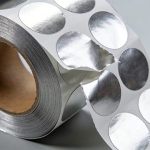 A roll of pure aluminum foil self-adhesive labels with metallic sheen, partially peeled to show the adhesive backing, on a clean industrial background.