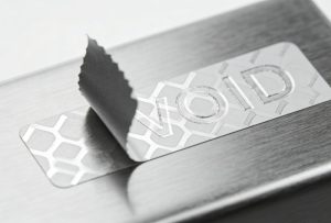 Close-up macro photo showing a tamper evident security label being removed, revealing a permanent "VOID" pattern left behind as tamper proof evidence.